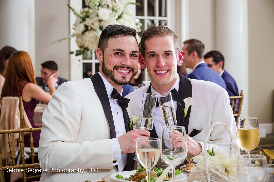 two grooms in white tuxedos black tie toasting with champagne at their wedding