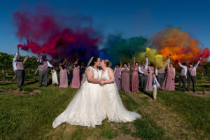 two brides kissing in front of wedding party holding color smoke bombs making a rainbow