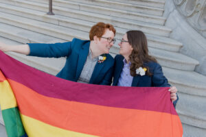 two brides, couple sitting on steps of library of congress holding gay flag smiling at each other