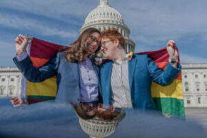 two brides with foreheads leaned in, smiling holding gay flag in washington dc